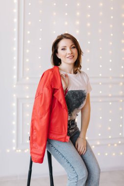 Beyaz tişörtlü, kot pantolonlu, kırmızı ceketli ve topuklu ayakkabılı şık genç kadın kırmızı merdivende oturuyor. stüdyo kısa.