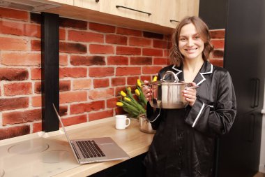 Çevrimiçi aşçılık dersi. Modern mutfakta elinde tencereyle blog yazarken dizüstü bilgisayar kullanan bir kadının kesilmiş fotoğrafı..