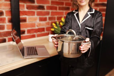 Çevrimiçi aşçılık dersi. Modern mutfakta elinde tencereyle blog yazarken dizüstü bilgisayar kullanan bir kadının kesilmiş fotoğrafı..