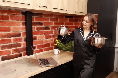 Çevrimiçi aşçılık dersi. Modern mutfakta elinde tencereyle blog yazarken dizüstü bilgisayar kullanan bir kadının kesilmiş fotoğrafı..