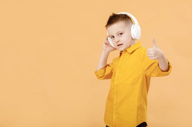 Sarı tişörtlü, kulaklıklı, komik, zeki okul çocuğunun portresi. Sarı stüdyo geçmişi. Eğitim. Bakıyor, gülümsüyor ve kameraya başparmağını gösteriyor. Yüksek kalite fotoğraf.