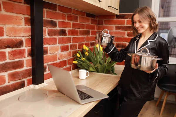 Çevrimiçi aşçılık dersi. Modern mutfakta elinde tencereyle blog yazarken dizüstü bilgisayar kullanan bir kadının kesilmiş fotoğrafı..