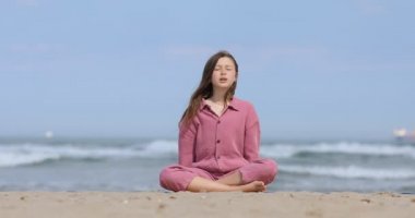 Çocuk plajda yoga egzersizi yapıyor. Pembe tulumlu, gözleri kapalı çocuk nilüfer çiçeği pozunda oturuyor ve kollarını namaste ile kavuşturuyor.