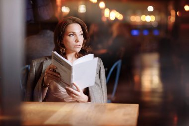 Genç ve güzel bir kadının portresi alacakaranlıkta kafede kağıt kitap okuyor. Kız çevrimiçi eğitim, uzaktan çalışma