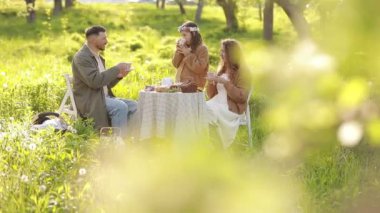 Tatilde mutlu bir aile. Anne, baba ve küçük kız yaz günbatımında yeşil parkta akşam yemeği partisinin tadını çıkarıyorlar. Anneler, babalar ve bebekler günü