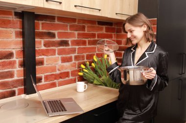 Çevrimiçi aşçılık dersi. Modern mutfakta elinde tencereyle blog yazarken dizüstü bilgisayar kullanan bir kadının kesilmiş fotoğrafı..