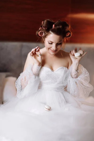 Beauty portrait of a beautiful smiling young brunette woman with bright pin-up make-up eating delicious french macaroons or macarons. girl in white dress and pink curlers sitting on the bed and relax.