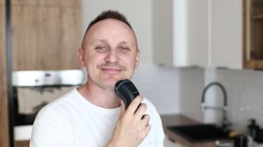 Beyaz tişörtlü komik beyaz erkek jiletle tıraş oluyor mutfakta tıraş oluyor, evde dans ediyor, mizahi müzik yapıyor.