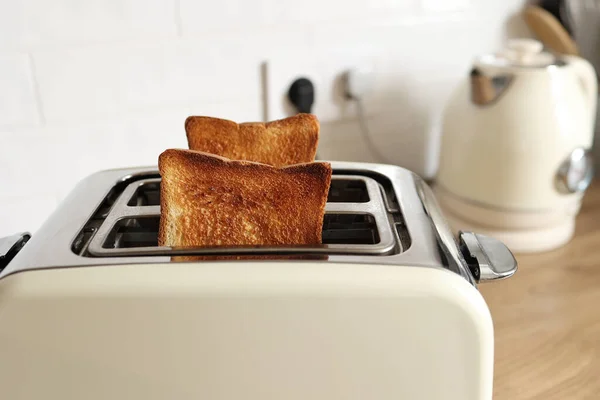Modern white toaster and roasted bread slices toasts inside on wooden table in kitchen.