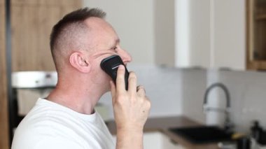Beyaz tişörtlü neşeli, komik, beyaz beyaz adam tıraş olurken tıraş olurken tıraş olurken, mutfakta tıraş olurken, evde müzik eşliğinde espri yaparken eğleniyor. Eğleniyorum. günlük yaşam.