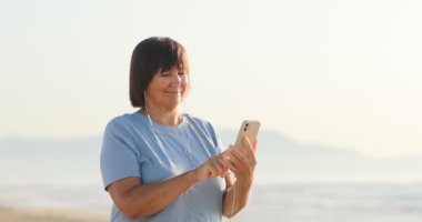 Mutlu son sınıf öğrencisi 50-60 yıl cep telefonu kullandı ve güneşli bir günde sahilde kulaklıkla müzik dinledi.