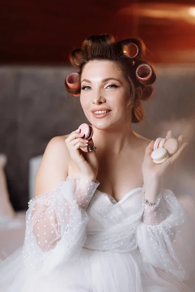 Beauty portrait of a beautiful smiling young brunette woman with bright pin-up make-up eating delicious french macaroons or macarons. girl in white dress and pink curlers sitting on the bed and relax.