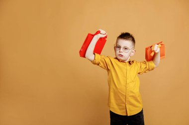 Elinde hediyeler ya da sarı arka planda alışveriş torbaları olan gülümseyen bir çocuk resmi. Gözlüklü çocuk paketleri ağırlık kaldırır gibi kaldırır..