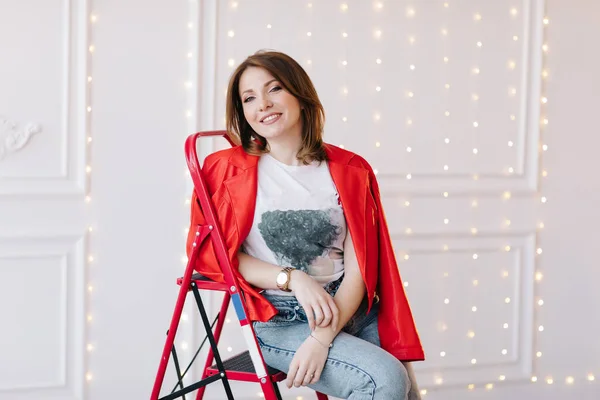 stylish young woman in white t shirt, jeans and red jacket and shoes on high heels is sitting on red ladder. studio short.