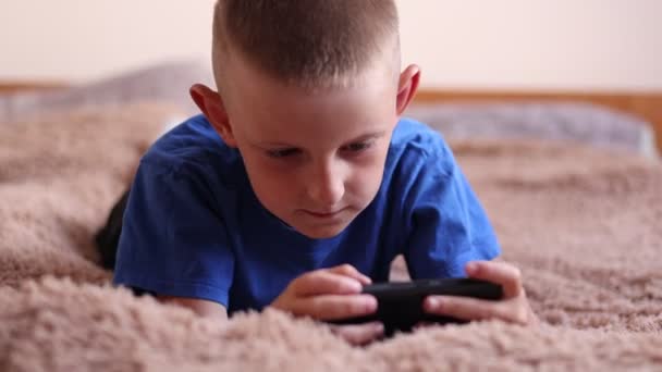 子供のモバイルデバイスのテーマへの依存 子供はベッドに横たわっていて スマートフォンでオンラインでプレイしている間に怒っています 積極的に行動する 子供のオンラインゲームへの中毒 仮想生活 — ストック動画