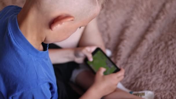 自宅の就学前の少年は スマートフォンを使用して ベッドの上に横たわっています 子供が携帯電話でビデオゲームをプレイ インターネットを閲覧 コンテンツを表示 ビデオ 肩越しにウィットカメラを撃つ — ストック動画