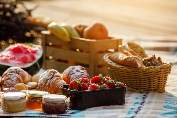 summer picnic basket with fruit and bakery on a blanket outdoors. Lunch with croissants, jam, watermelon, strawberry and fresh fruits in wooden box in the park. Copy space