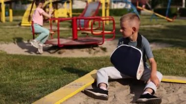 Kırık kollu ağlayan bir çocuk dışarıda oturur ve çocuk parkında nasıl oynadığına bakar. Sağlık kazası ve tıbbi kaza kavramı. En kötü yaz tatili.