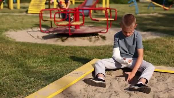 腕の骨折で泣いている子供は屋外に座って子供の女の子が遊び場で遊んでいる様子を見ています 医療事故という概念です 最悪の夏休み — ストック動画