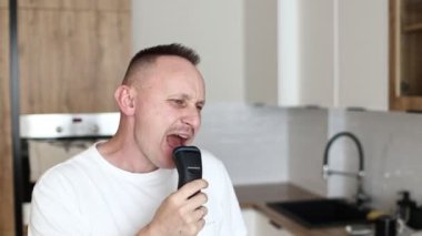 Beyaz tişörtlü neşeli, komik, beyaz beyaz adam tıraş olurken tıraş olurken tıraş olurken, mutfakta tıraş olurken, evde müzik eşliğinde espri yaparken eğleniyor. Eğleniyorum. günlük yaşam.