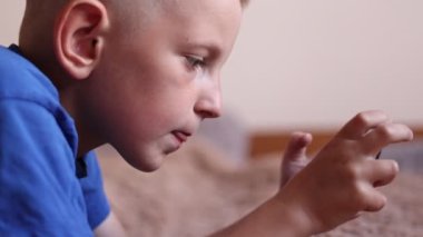 Anaokulundaki çocuk yatağında uzanmış, akıllı telefon kullanıyor. Cep telefonuyla video oyunu oynayan çocuk, İnternet, Görüntü, Videolar. Omzunda kamera var..