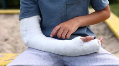 Açık havada kırık bir bacağı olan küçük bir çocuk. Bir çocuğun kolu kırılmış. Yaz tatilinde kırık yaralanması. Çocuk kolundaki alçıya dokunuyor.