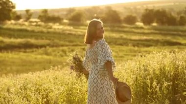 Bir buket yabani çiçekle yaz çayırında daireler çizen mutlu genç bir kadın. Hasır şapkalı kız güneşli bir günde yeşil tarlada yürüyor. Aktif yaşam tarzı. Güzel, neşeli kız tarlada yürüyor.