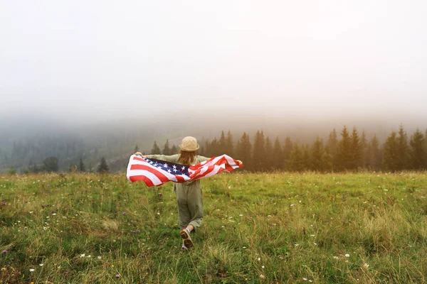 Beautiful child girl in hat with the American flag on foggy mountains. Independence Day of United States USA on 4th of July with family. Freedom. American patriotic holiday concept.