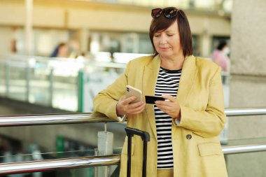 Yetişkin, güzel bir kadın akıllı telefon ve kredi kartını online alışveriş için kullanıyor. Bavullu bayan havaalanında, iş ve seyahat konseptinde bekliyor..