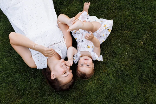Young beautiful mother and baby girl playing and having fun on grass at summer. mom and child are lying down on ground outdoors. Parent with kid spend time together. healthy family. Positive emotions