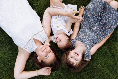 Aile günü. Güzel genç anne ve iki küçük kızı parkta dışarıda oynuyorlar. Anne ve çocuklar birlikte yeşil çimlerin üzerinde yatıp dinleniyorlar. Anneler Günü. Üst Manzara. Mutlu çocukluk