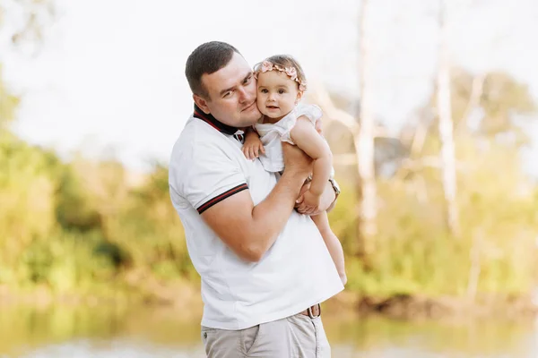 Father\'s day. Dad is hugging his baby daughter outdoors and having fun by the pond. Family enjoying life together on nature. Happy young family day. The concept of family summer holiday