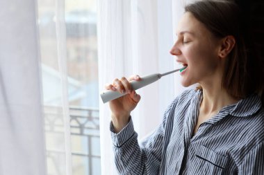 Kız dişlerini elektrikli fırçayla fırçalıyor. Genç ve güzel bir kız dişlerini aynanın önünde elektrikli bir diş fırçasıyla fırçalıyor. Ağız temizliği.