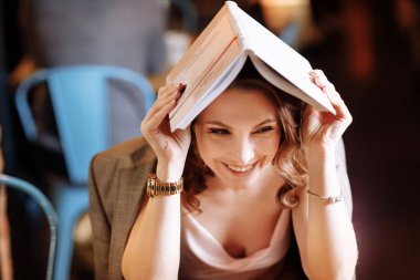 portrait of young beautiful woman is reading paper book in cafe in dusk. girl is online study, remote work