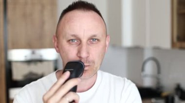 Beyaz tişörtlü neşeli, komik, beyaz beyaz adam tıraş olurken tıraş olurken tıraş olurken, mutfakta tıraş olurken, evde müzik eşliğinde espri yaparken eğleniyor. Eğleniyorum. günlük yaşam.
