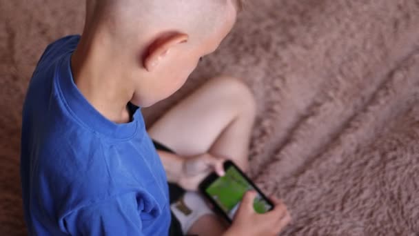 自宅の就学前の少年は スマートフォンを使用して ベッドの上に横たわっています 子供が携帯電話でビデオゲームをプレイ インターネットを閲覧 コンテンツを表示 ビデオ 肩越しにウィットカメラを撃つ — ストック動画