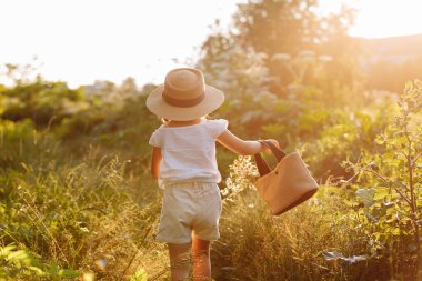 Little girl walks in the rays of a sunset in a flowering meadow, enjoying the summer, warmth, flowers, freedom. Child with straw hat and bag is having fun outdoors. back view.