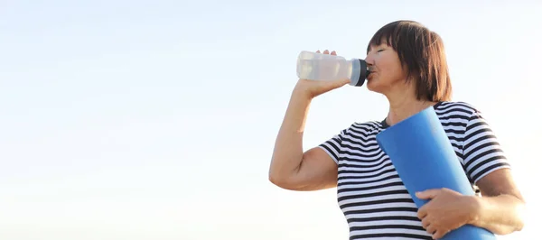 banner of mature woman drinking water from bottle after fitness exercises or yoga outdoors in morning on summer. Fitness female profile on blue sky background, concept of sport and relaxation