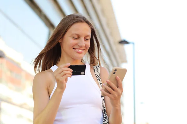 Happy smile young adult woman consumer using credit card and smartphone. people shopping via online application media concept. big shopping mall on summer day background with copy space.