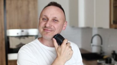 Beyaz tişörtlü komik beyaz erkek jiletle tıraş oluyor mutfakta tıraş oluyor, evde dans ediyor, mizahi müzik yapıyor.