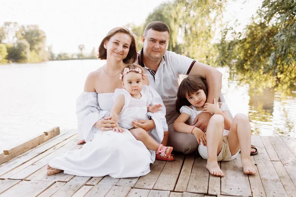 Happy young family near lake, pond on summer. Mother, father and two child daughter smiling while spending free time outdoors. Family enjoying life together. People having fun in nature. Family day.