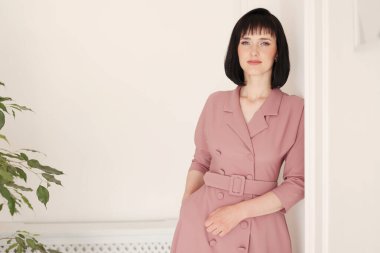 Çekici, gülümseyen esmer ve kısa saçlı bir kadının portresi. Beyaz, şık bir duvar arkasında yeşil çiçekli yalnız kalıyor. Yumuşak pembe elbiseli kadın kameraya bakıyor..