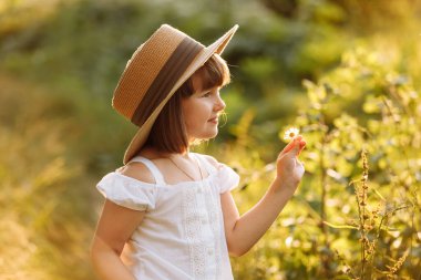 Portrait of cute little girl is smelling wild flower and walks in the rays of a sunset in a flowering meadow, enjoying the summer, warmth, freedom. Child in mommys big straw hat having fun outdoors.