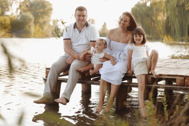 Family group active having fun by the lake. Young mother, father and two cute child daughter girls sitting on wooden bridge and splashing water with their legs by the pond. Happy family day