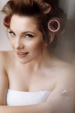 Güzellik konsepti Beyaz battaniye ve pembe bukleli parlak makyajlı, gösterişli, genç bir esmer kadın. Kız pencerenin kenarında oturuyor ve kameraya bakıyor. Uluslararası Kadınlar Günü.