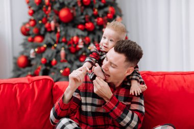 young caucasian father with baby boy are having fun on red sofa by the Christmas tree at home. Dad and son with the same pyjamas celebrating Xmas and New Years family holiday vacation. Fathers day.
