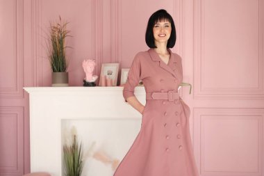 Çekici, gülümseyen esmer ve kısa saçlı kadının portresi kollarını kavuşturmuş ve iç mekanda tek başına kalıyordu. Yumuşak pembe elbiseli kadın kameraya bakıyor..