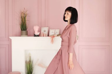 Çekici, gülümseyen esmer ve kısa saçlı kadının portresi kollarını kavuşturmuş ve iç mekanda tek başına kalıyordu. Yumuşak pembe elbiseli kadın kameraya bakıyor..