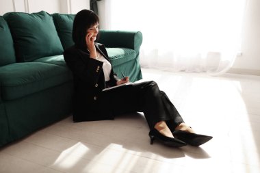 İş kadını akıllı bir telefondan konuşuyor. Yorgun esmer kadın iş elbisesi ve yüksek topuklu ayakkabılarıyla yeşil kanepenin yanında oturuyor. İç kısımda yeşil bitkiler var. Evden çalış.