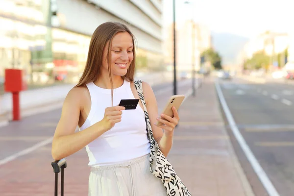 Beautiful young smiling woman with smartphone and credit card by the airport. Happy girl with suitcase after arrived airplane booking and paying for hotel. summer vacation. new abroad journey travel.